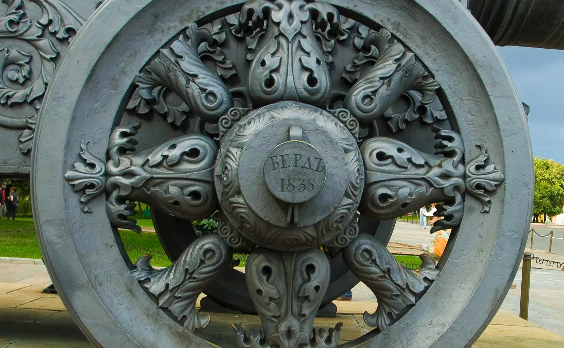 Canhao monumental de bronze historico em tom esverdeado montado sobre uma base de quatro rodas de metal esculpido no Kremlin de Moscou. A enorme boca redonda do cano aponta para o alto em direcao a direita, diante da qual repousam tres imensas esferas de ferro preto em formato de piramide cercadas por uma corrente de protecao. Ao fundo destacam-se arvores verdes e a fachada clara de um edificio historico sob o ceu azul ensolarado.