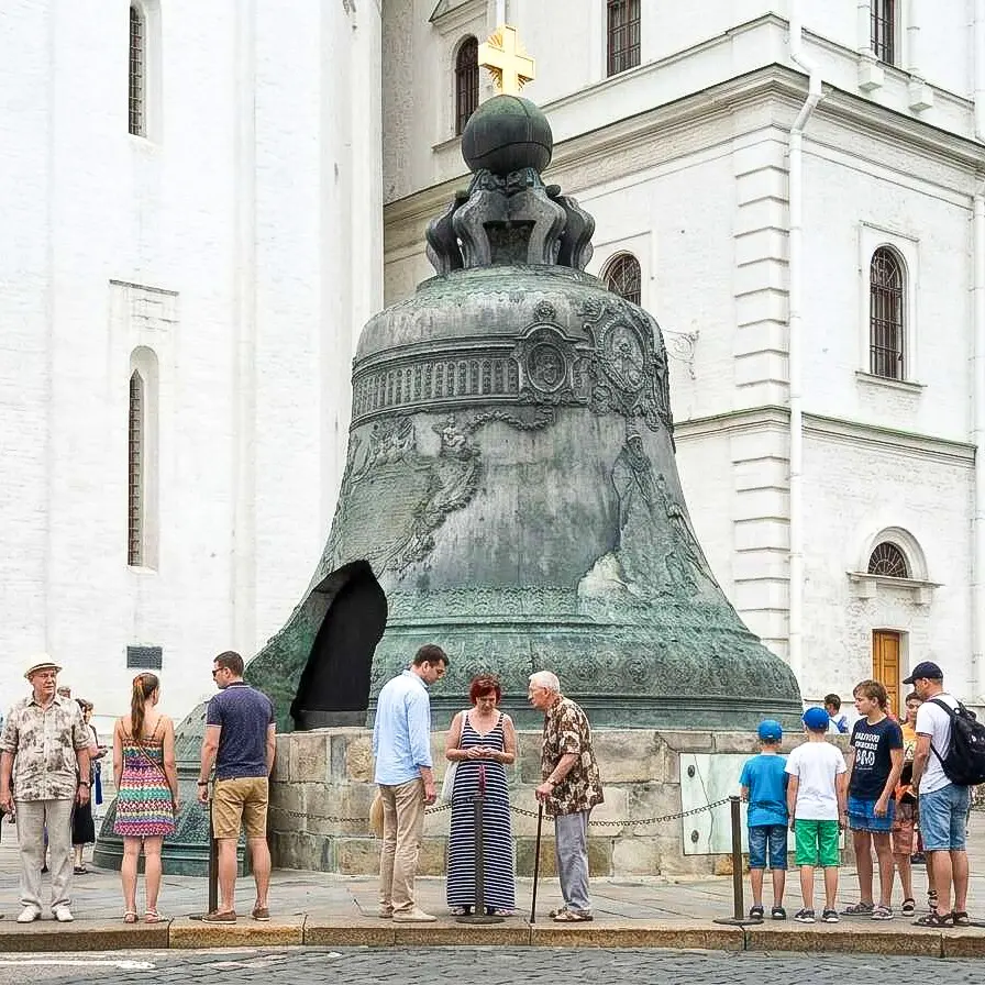 Sino monumental de bronze em tom cinza-esverdeado montado sobre um pedestal circular de pedra no Kremlin de Moscou, diante da parede de um predio historico branco. Na base da peca ha uma enorme abertura triangular, ao lado da qual repousa verticalmente o bloco quebrado de metal da altura de uma pessoa adulta, cercado por visitantes na calcada.