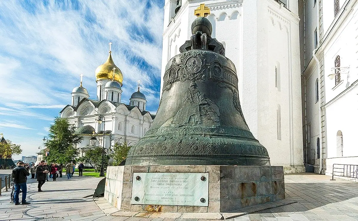 Sino monumental de bronze em tom cinza-esverdeado montado sobre um pedestal circular de pedra no Kremlin de Moscou, diante da parede de um predio historico branco. Na base da peca ha uma enorme abertura triangular, ao lado da qual repousa verticalmente o bloco quebrado de metal da altura de uma pessoa adulta, cercado por visitantes na calcada.