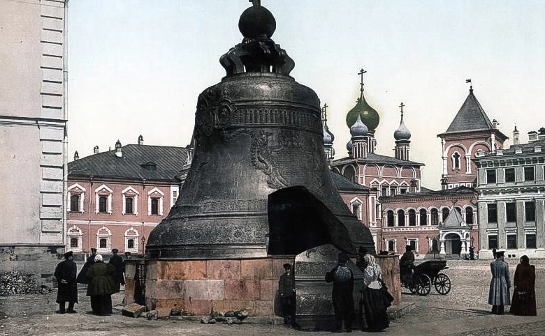 Sino monumental de bronze em tom cinza-esverdeado montado sobre um pedestal circular de pedra no Kremlin de Moscou, diante da parede de um predio historico branco. Na base da peca ha uma enorme abertura triangular, ao lado da qual repousa verticalmente o bloco quebrado de metal da altura de uma pessoa adulta, cercado por visitantes na calcada.