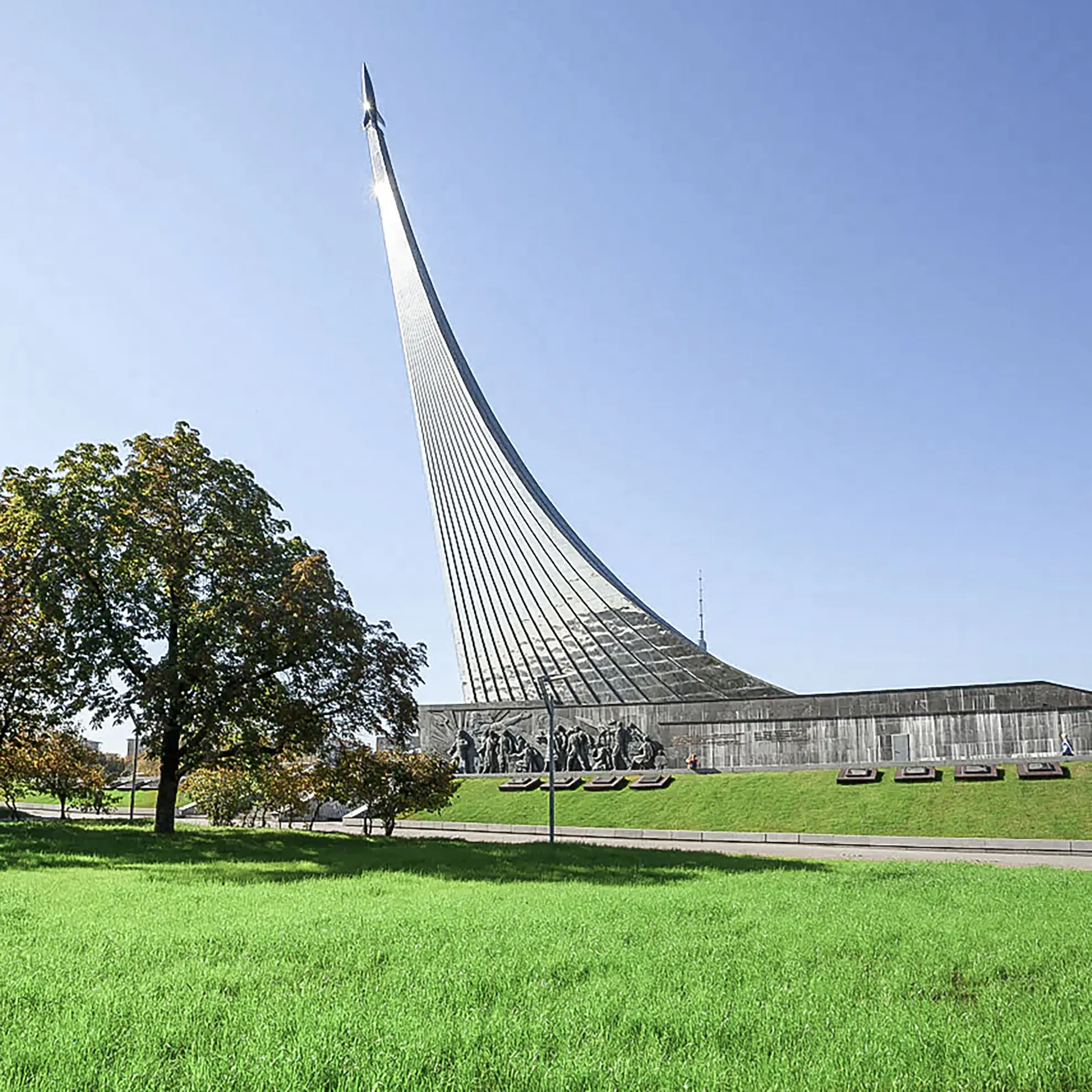 Obelisco monumental de titânio reproduzindo a cauda de fumaça de um foguete que sobe em direção ao céu azul limpo. No topo da lâmina metálica curvada destaca-se o foguete prateado, apoiado sobre uma base retangular de pedra escura ladeada por árvores e um amplo gramado verde.