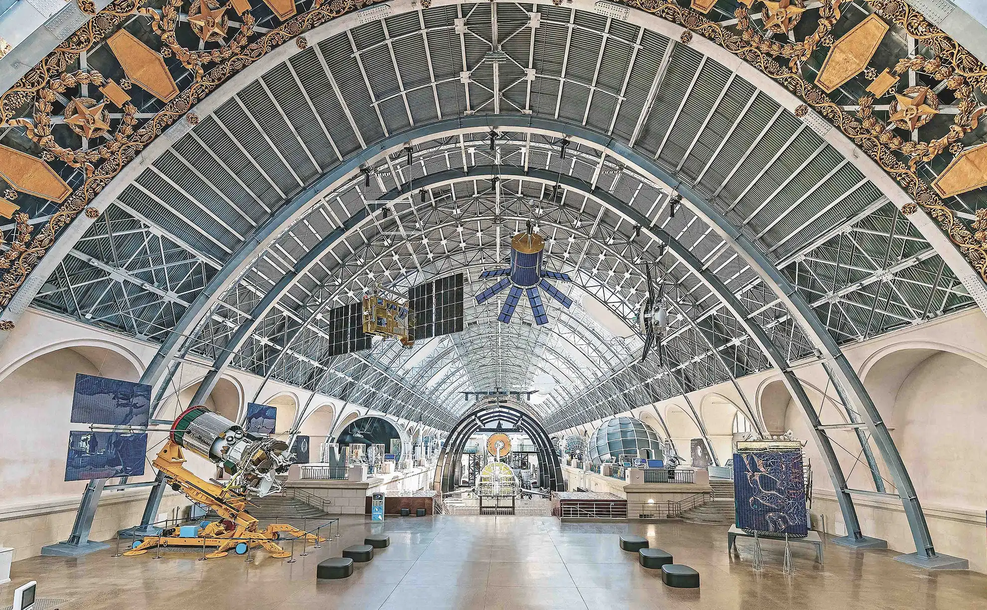 Réplica de 38 metros do foguete espacial Vostok em rampa de lançamento diante da fachada com cúpula do pavilhão Cosmos na praça da VDNKh, com pedestres caminhando e esculturas douradas no topo.