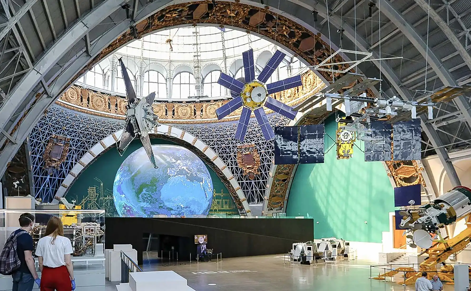 Réplica de 38 metros do foguete espacial Vostok em rampa de lançamento diante da fachada com cúpula do pavilhão Cosmos na praça da VDNKh, com pedestres caminhando e esculturas douradas no topo.