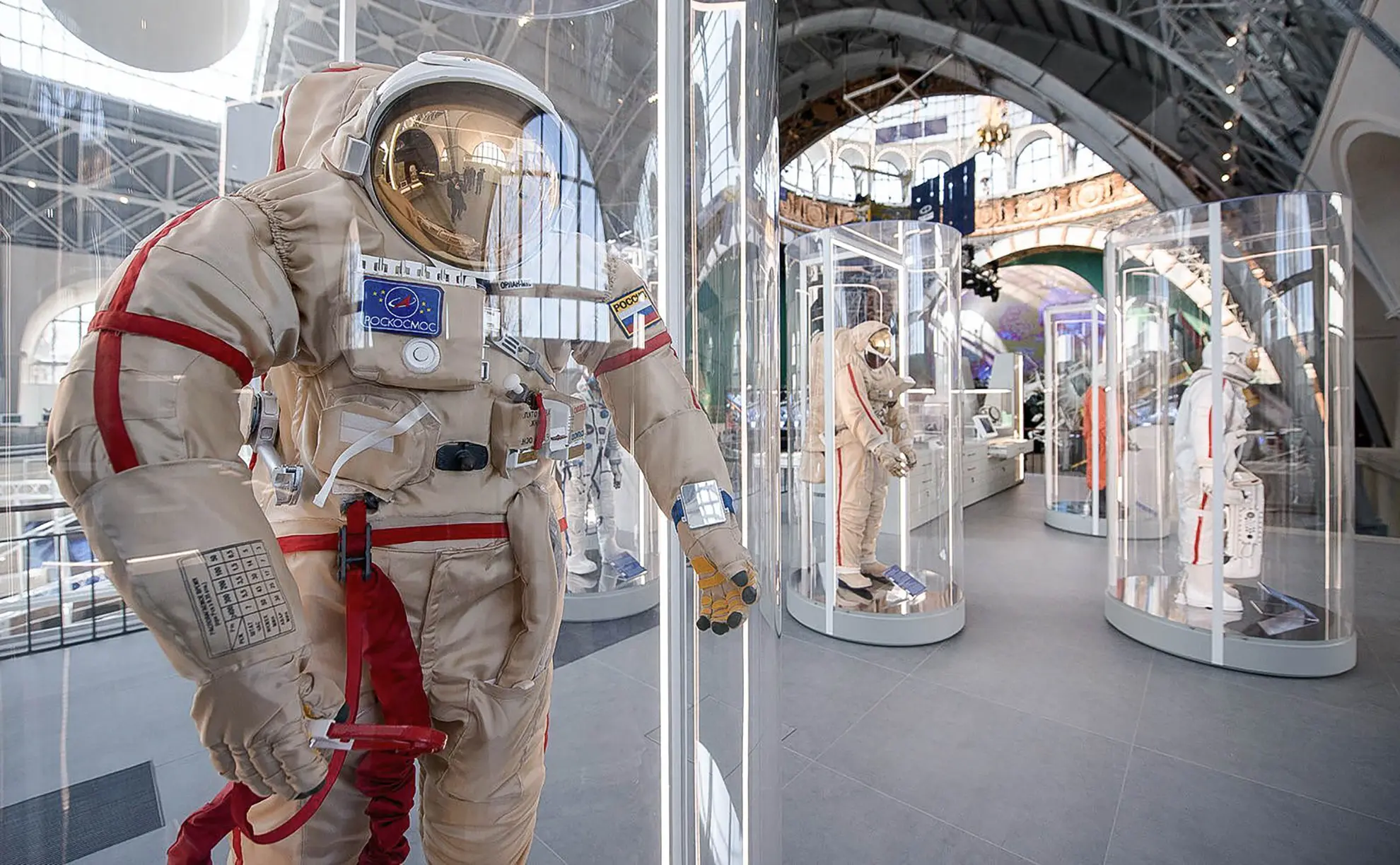 Traje espacial branco modelo Orlan com visor dourado exposto em uma coluna de vidro transparente no interior do pavilhão de exposições aeroespaciais.