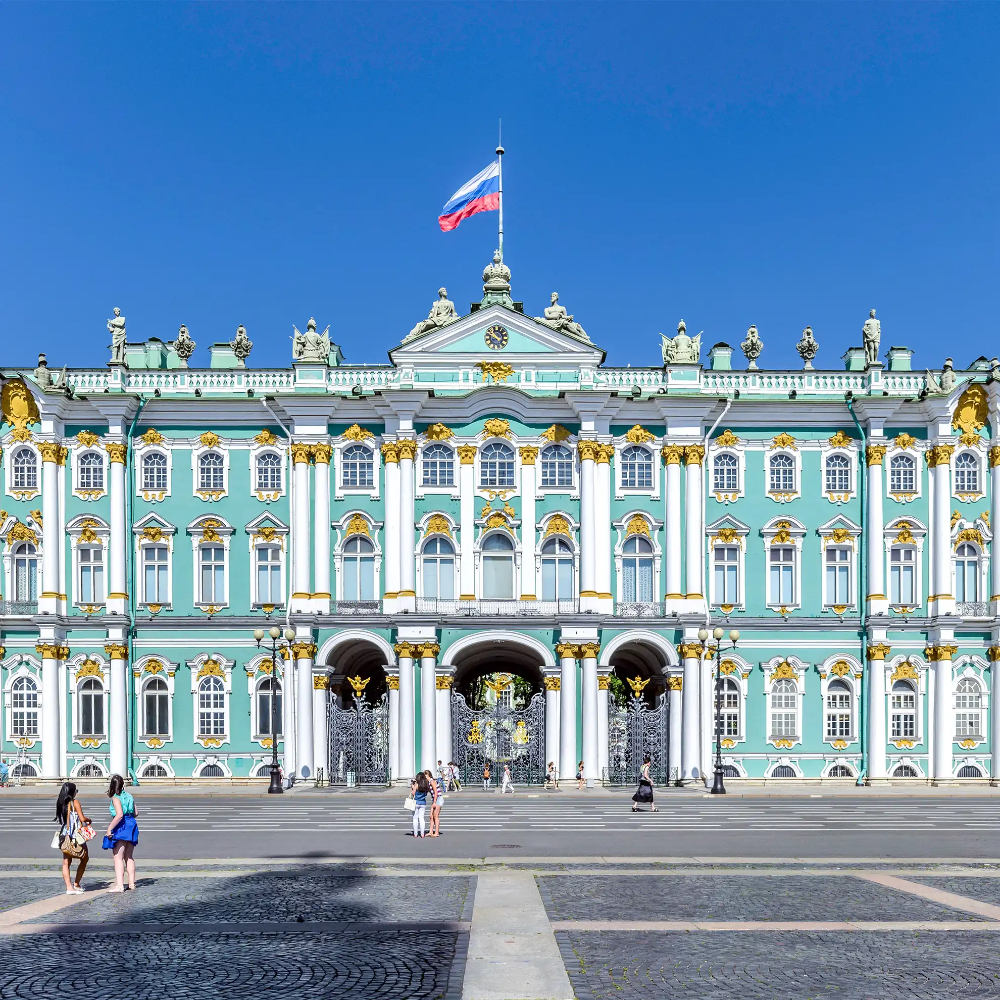 Fachada imponente em verde-turquesa do Palácio de Inverno em São Petersburgo sob céu azul límpido. O palácio barroco de três pavimentos exibe colunas brancas com detalhes dourados, molduras curvas nas janelas e dezenas de estátuas na cornija superior, destacando-se a bandeira russa ao centro e a ampla esplanada frontal de paralelepípedos com transeuntes.
