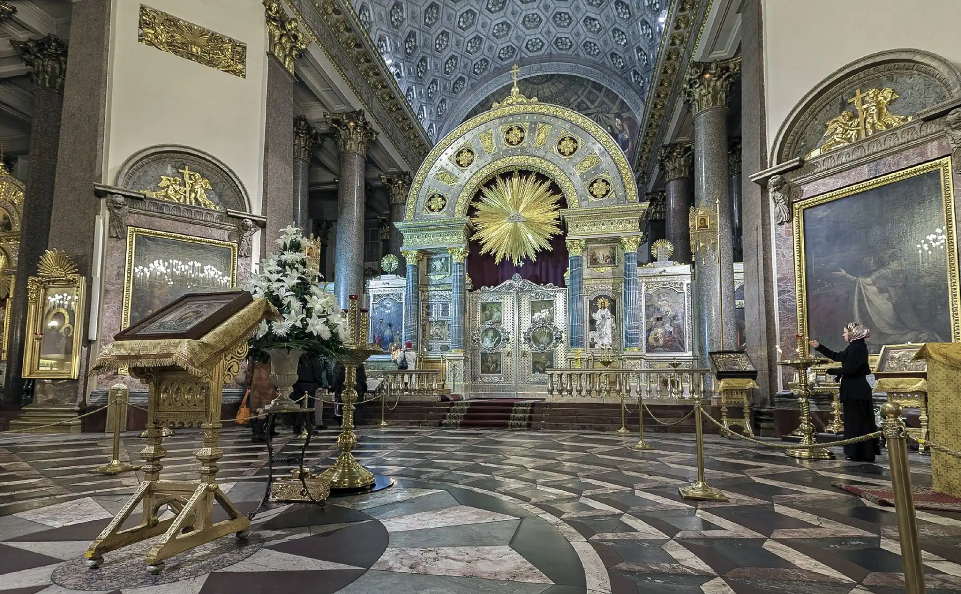 Vista panorâmica da Catedral de Kazan em São Petersburgo sob céu azul claro com nuvens dispersas. Diante da cúpula central ergue-se a monumental colunata em ferradura de pedra clara delimitada por pórticos clássicos nas pontas, cercada em primeiro plano por amplos gramados verdes com caminhos curvos, arbustos floridos, bancos e chafariz.