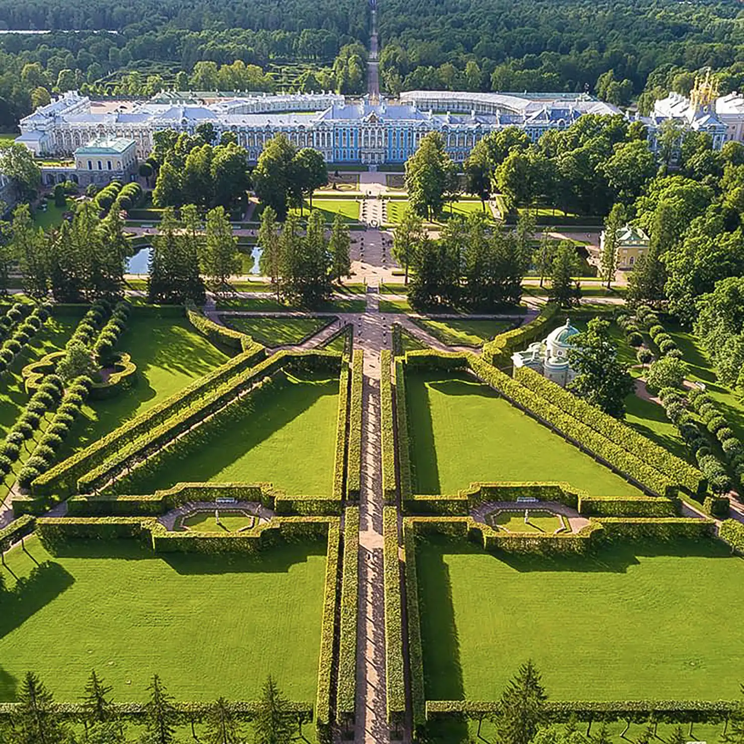 Vista aérea em plano geral do Palácio de Catarina e do parque histórico na Vila do Czar sob iluminação solar plena. Em primeiro plano destacam-se os gramados verdes recortados e sebes podadas em desenhos geométricos com caminhos retos, revelando ao fundo entre copas de árvores a longa fachada azul do palácio imperial com cúpulas douradas.