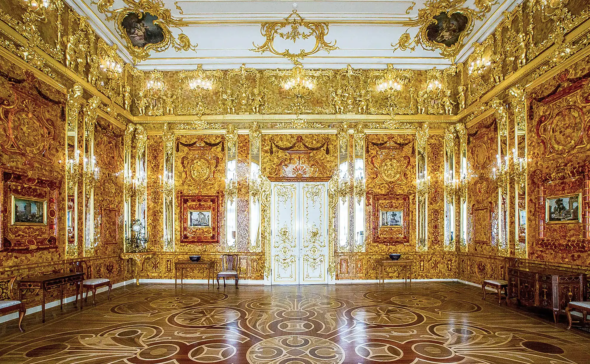 Vista aérea em plano geral do Palácio de Catarina e do parque histórico na Vila do Czar sob iluminação solar plena. Em primeiro plano destacam-se os gramados verdes recortados e sebes podadas em desenhos geométricos com caminhos retos, revelando ao fundo entre copas de árvores a longa fachada azul do palácio imperial com cúpulas douradas.