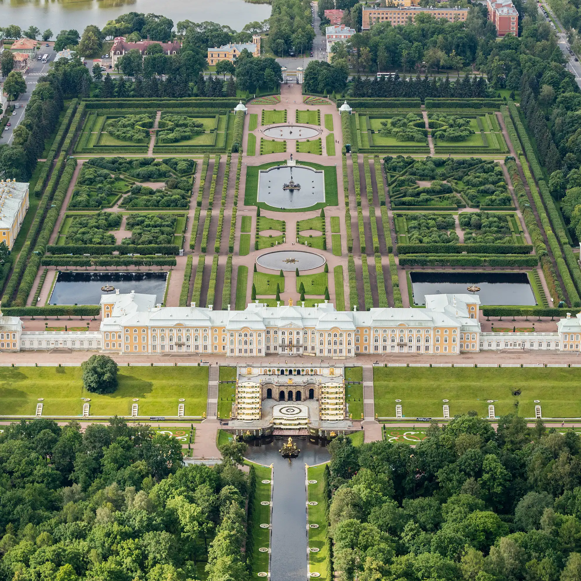 Vista aérea em plano geral do complexo imperial de Peterhof sob luz solar plena. Ao centro estende-se a fachada do Grande Palácio encimada por cúpulas douradas, ladeada pelo traçado geométrico do Jardim Superior com lagos de um lado e, do outro, pela descida em degraus da Grande Cascata até o canal marítimo cercado por bosques verdes.