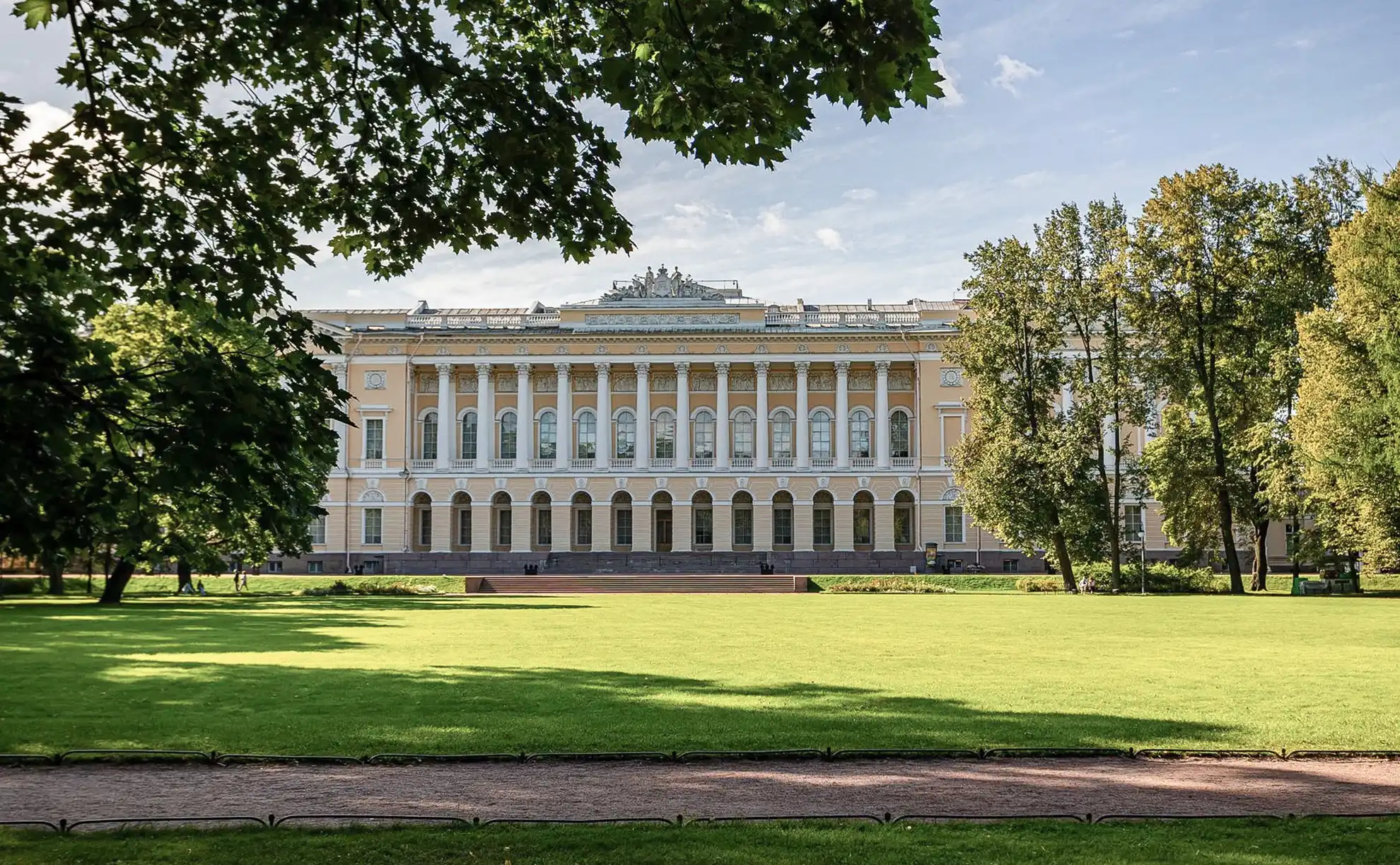 Ampla alameda de terra batida no Jardim Mikhailovsky sob iluminação solar plena em São Petersburgo. Árvores frondosas ladeiam o caminho projetando sombras sobre o solo claro, com bancos de ferro forjado e poste clássico à esquerda e gramado verde bem aparado estendendo-se à direita.