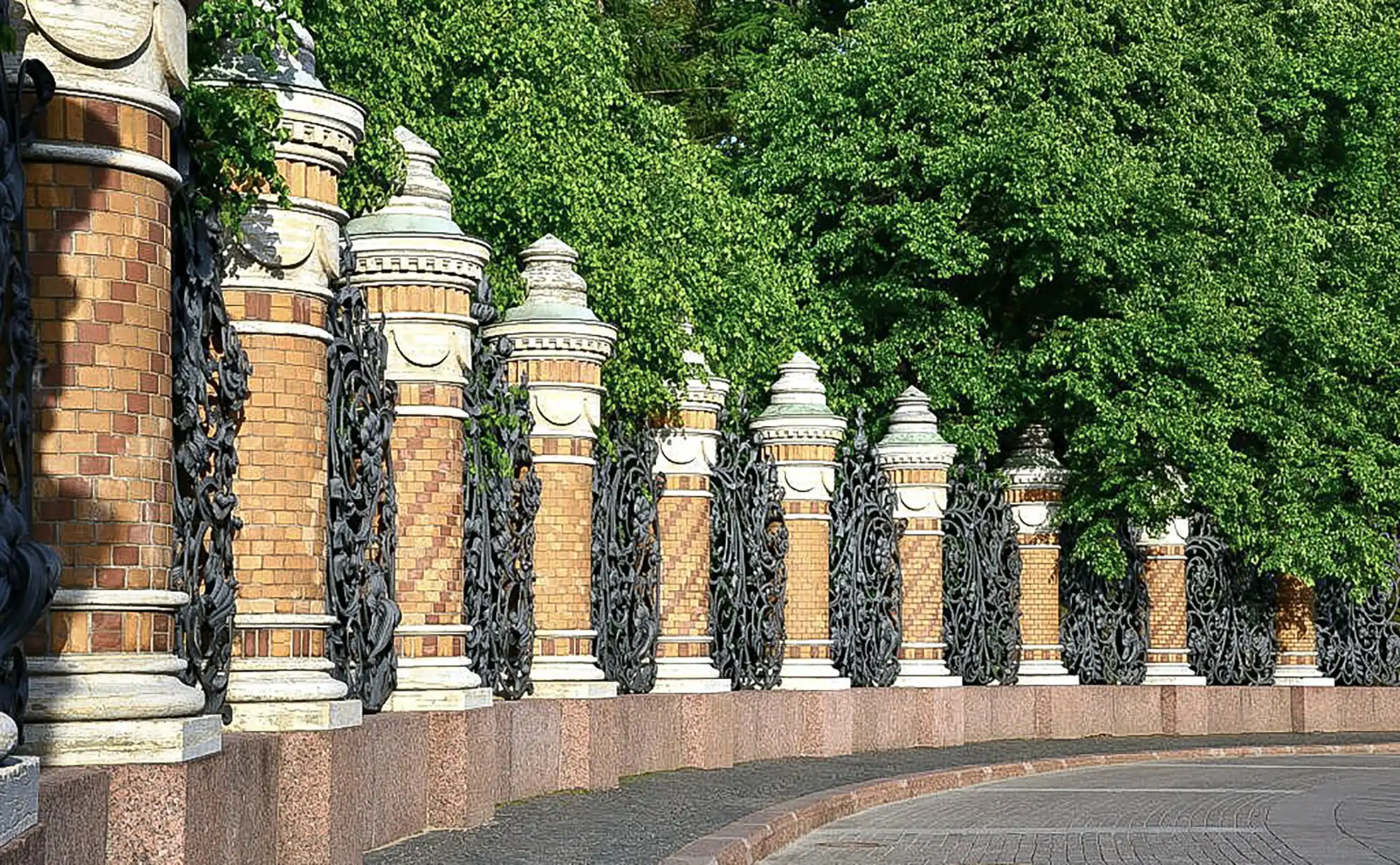 Ampla alameda de terra batida no Jardim Mikhailovsky sob iluminação solar plena em São Petersburgo. Árvores frondosas ladeiam o caminho projetando sombras sobre o solo claro, com bancos de ferro forjado e poste clássico à esquerda e gramado verde bem aparado estendendo-se à direita.