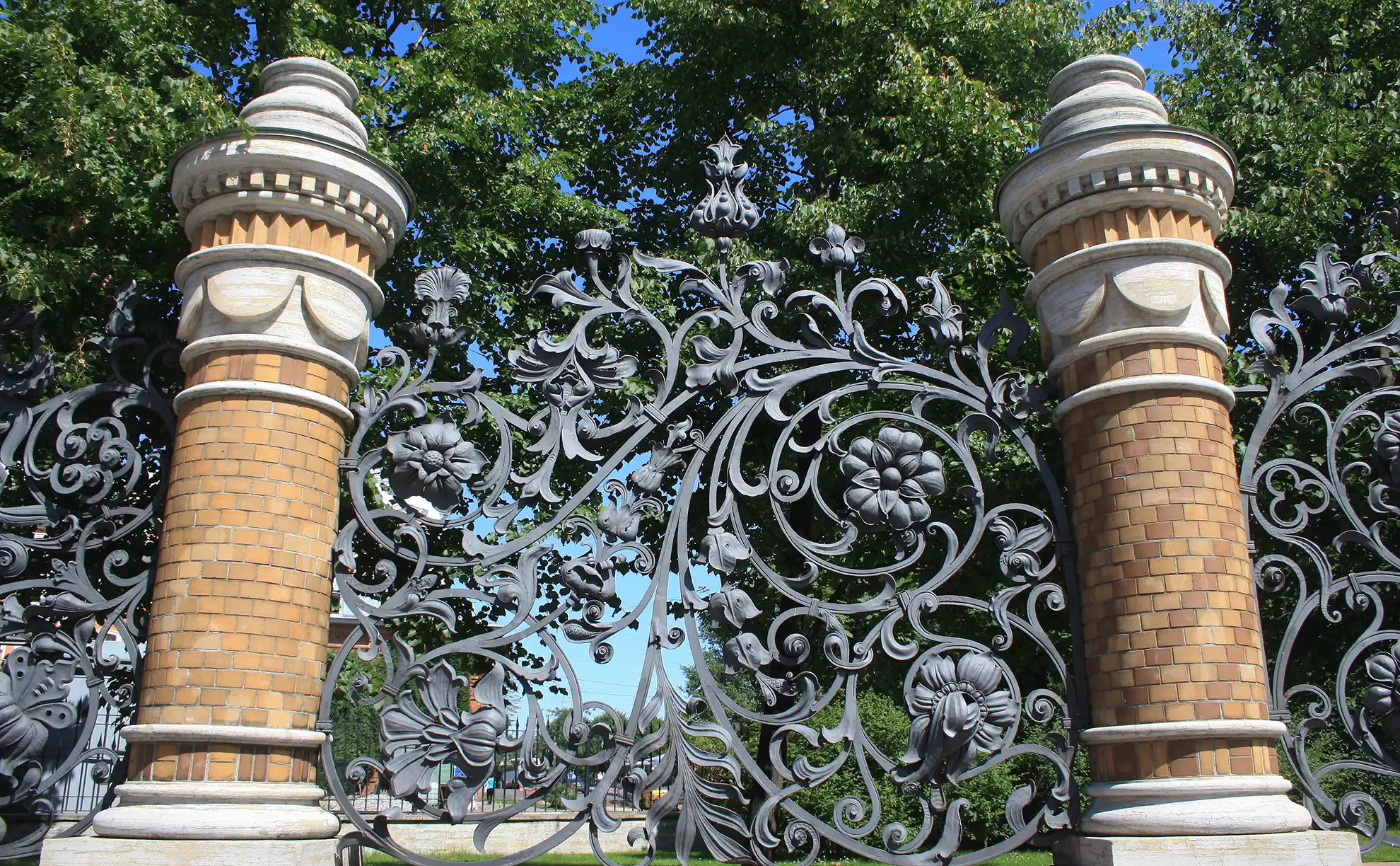 Detalhe em primeiro plano da grade de ferro forjado do Jardim Mikhailovsky ladeada por dois pilares de tijolos amarelados. O painel central exibe escultura metálica densa composta por folhas entrelaçadas e flores abertas em relevo contra o fundo de folhagens verdes e céu azul.