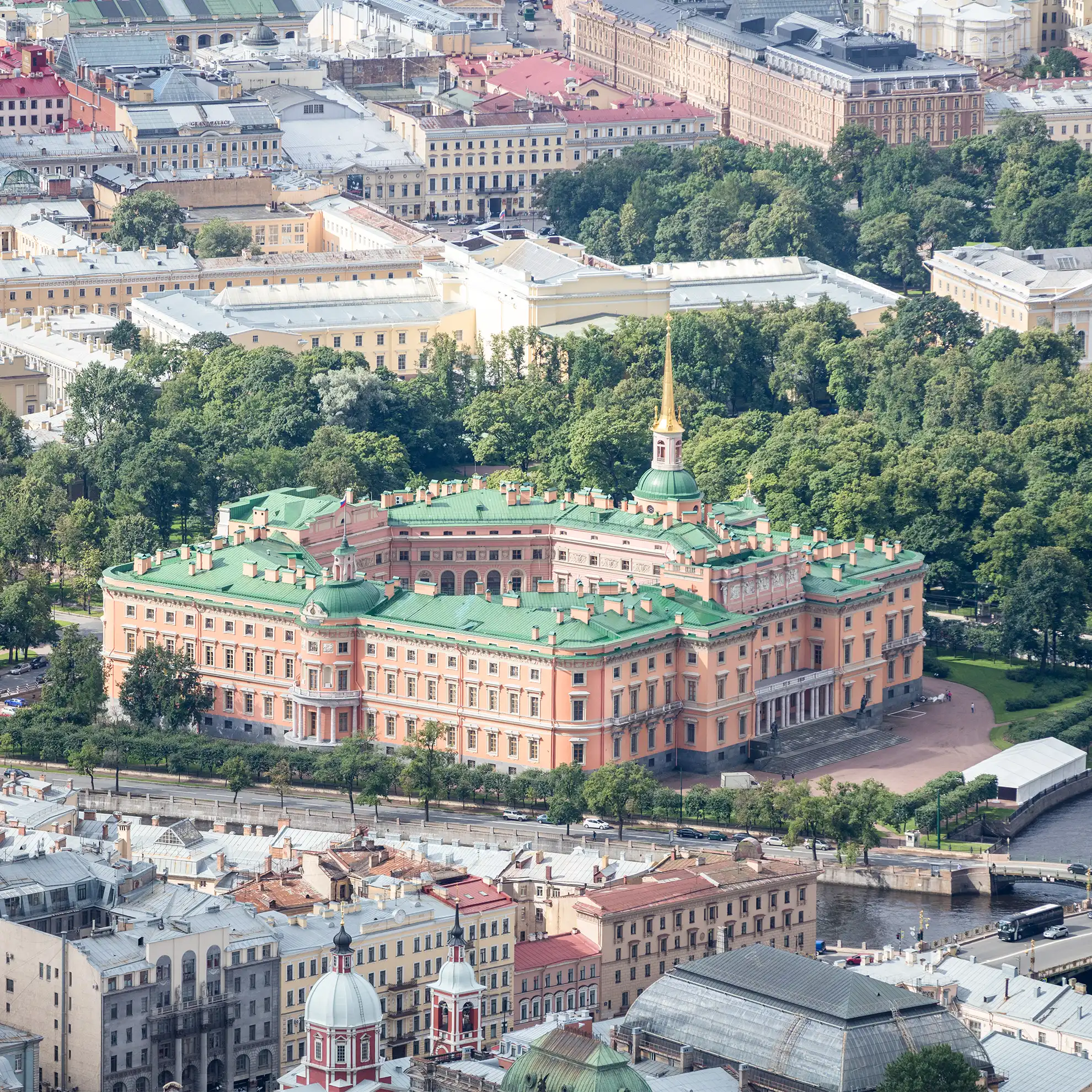 Vista aérea do Castelo Mikhailovsky em tom terracota destacando seu pátio octogonal interno e o pináculo dourado sob céu claro. O complexo fortificado ergue-se cercado pela arborização dos parques vizinhos e pelo casario histórico de São Petersburgo, margeado à direita pelas águas de canais urbanos cruzados por pontes de tráfego de veículos.