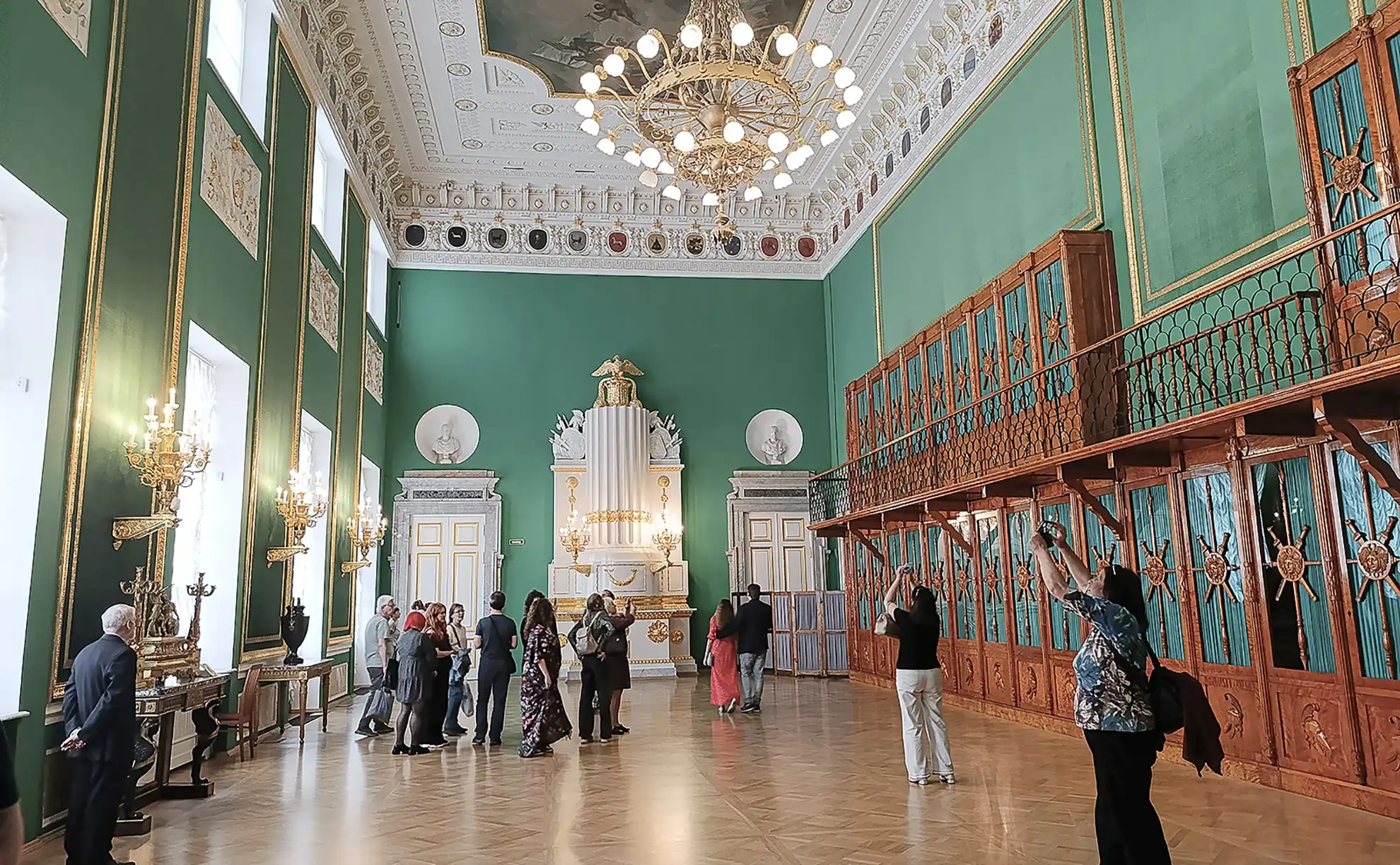 Amplo aposento do Corpo da Guarda no Castelo Mikhailovsky exibindo paredes altas em verde intenso e piso de marchetaria. O lado direito é dominado por uma estrutura de galeria em madeira escura de dois níveis destinada ao porte de armas, com estufa cerâmica branca ladeada por detalhes em ouro ao fundo sob lustre dourado suspenso.