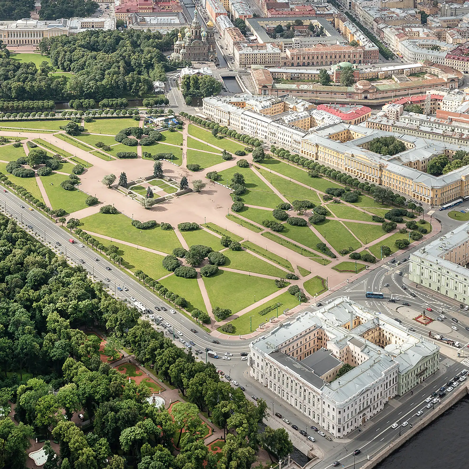 Vista aérea do Campo de Marte em São Petersburgo em plano aberto sob luz diurna. O amplo parque triangular é recortado por caminhos de pedestres e gramados verdes com grupos de árvores, ladeado à direita pelo casario clássico do centro histórico e na base pela orla fluvial e pontes com tráfego urbano.