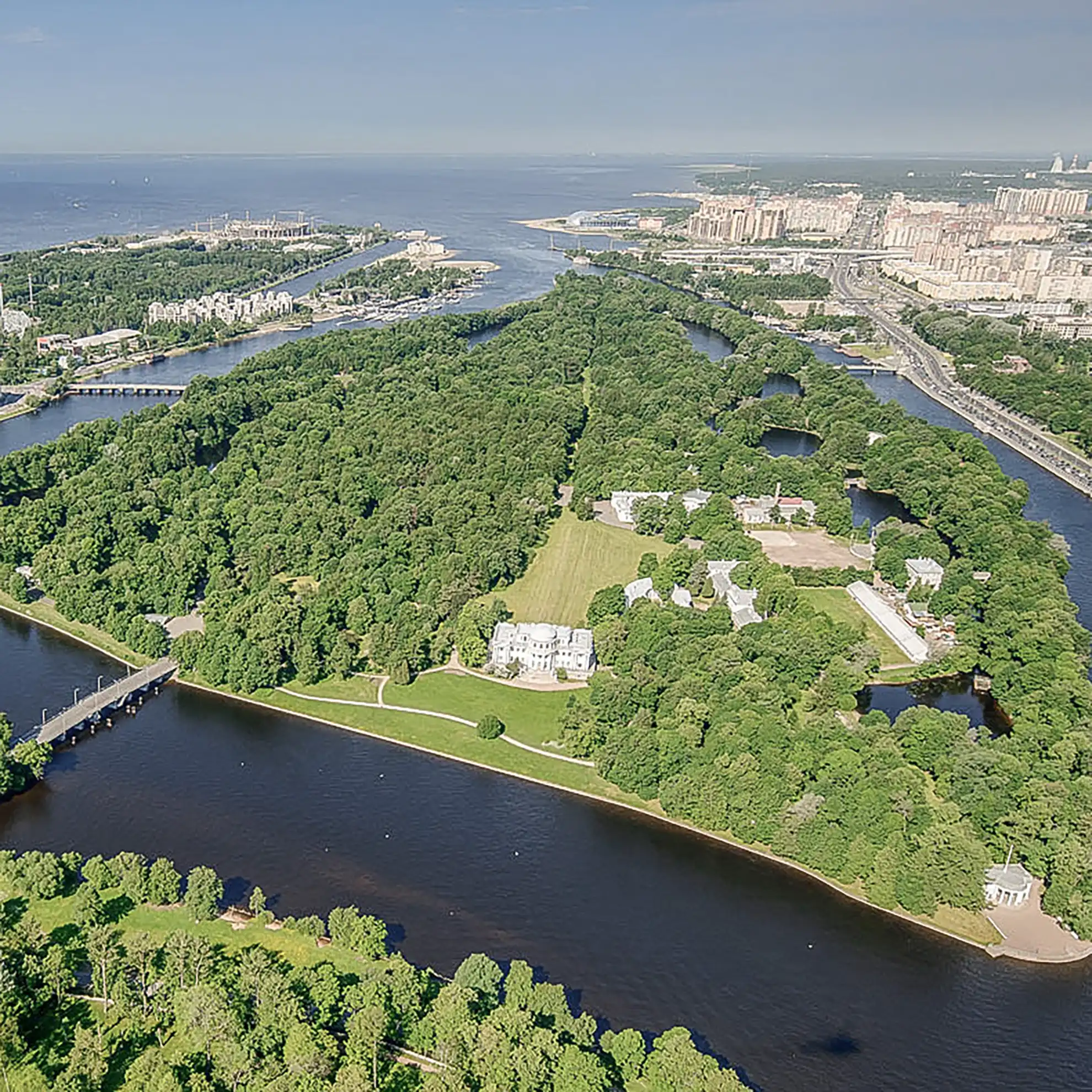 Vista aérea panorâmica da Ilha Yelagin em São Petersburgo sob dia ensolarado. A densa mancha verde de árvores é contornada pelas curvas largas do rio Neva, destacando ao centro a estrutura do Palácio Yelagin cercada por gramados abertos, com o Golfo da Finlândia e os bairros da cidade ao fundo.