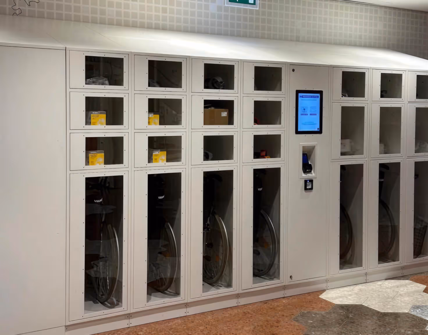White lockers with glass doors containing bicycles and equipment, featuring a touchscreen interface in the center.