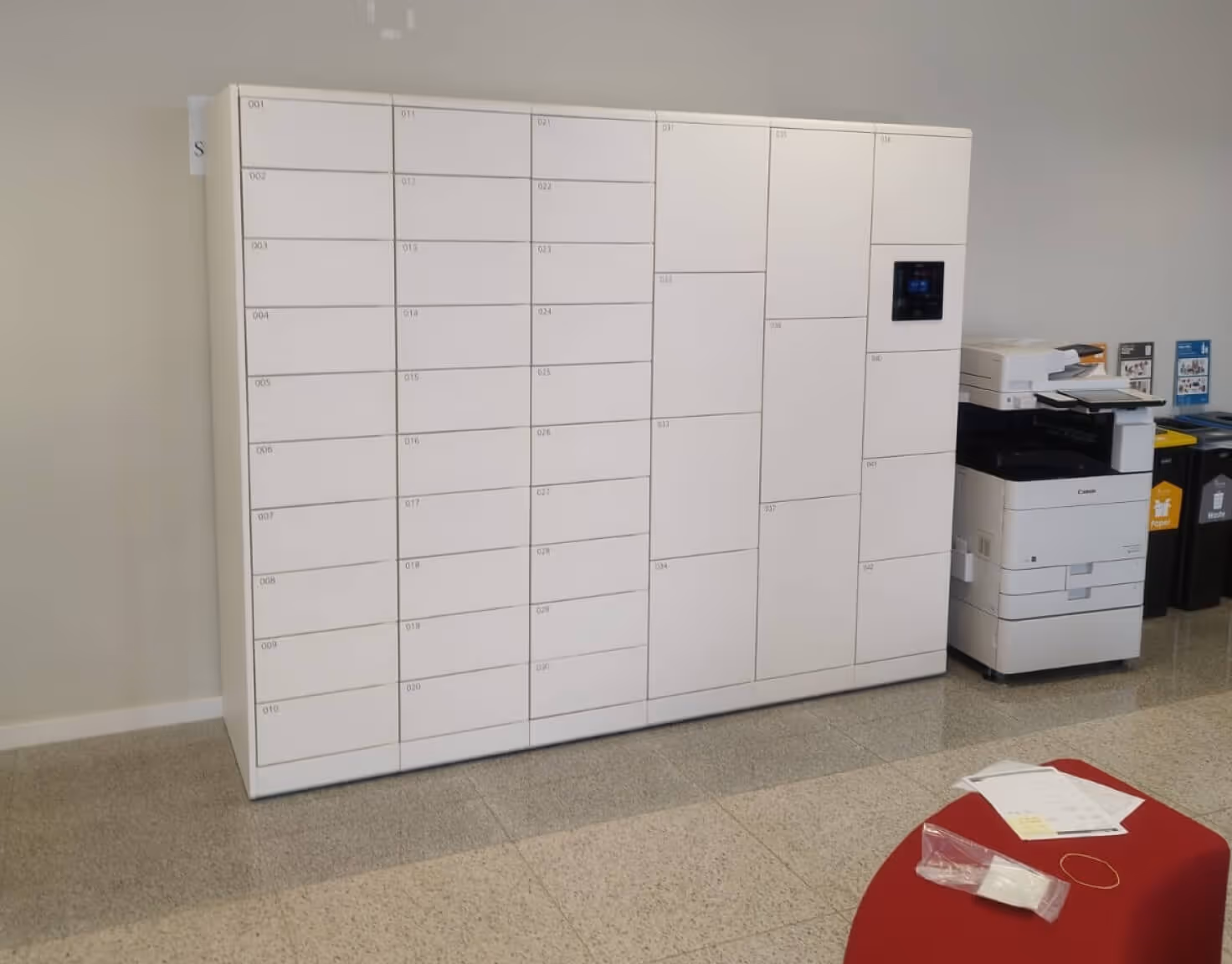 White parcel locker system with multiple numbered compartments next to a white Canon printer and recycling bins in an indoor office setting.
