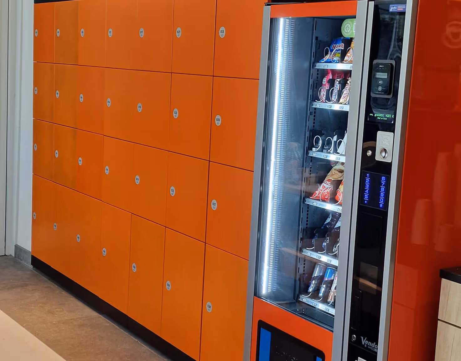 Orange smart locker system with multiple compartments next to a vending machine stocked with snacks.