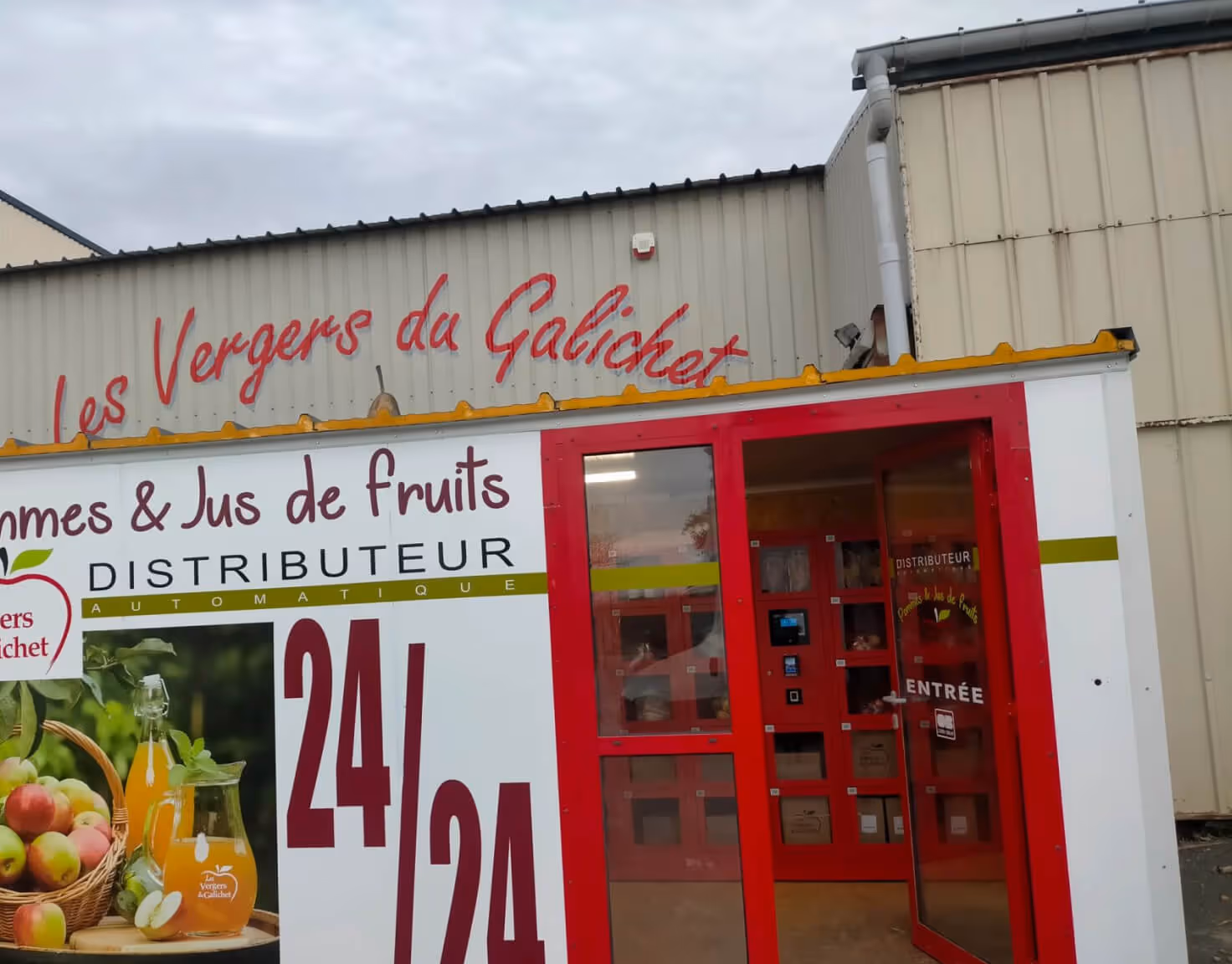 Automatic fruit and juice vending machine with red door and signage for Les Vergers du Galichet operating 24/7.
