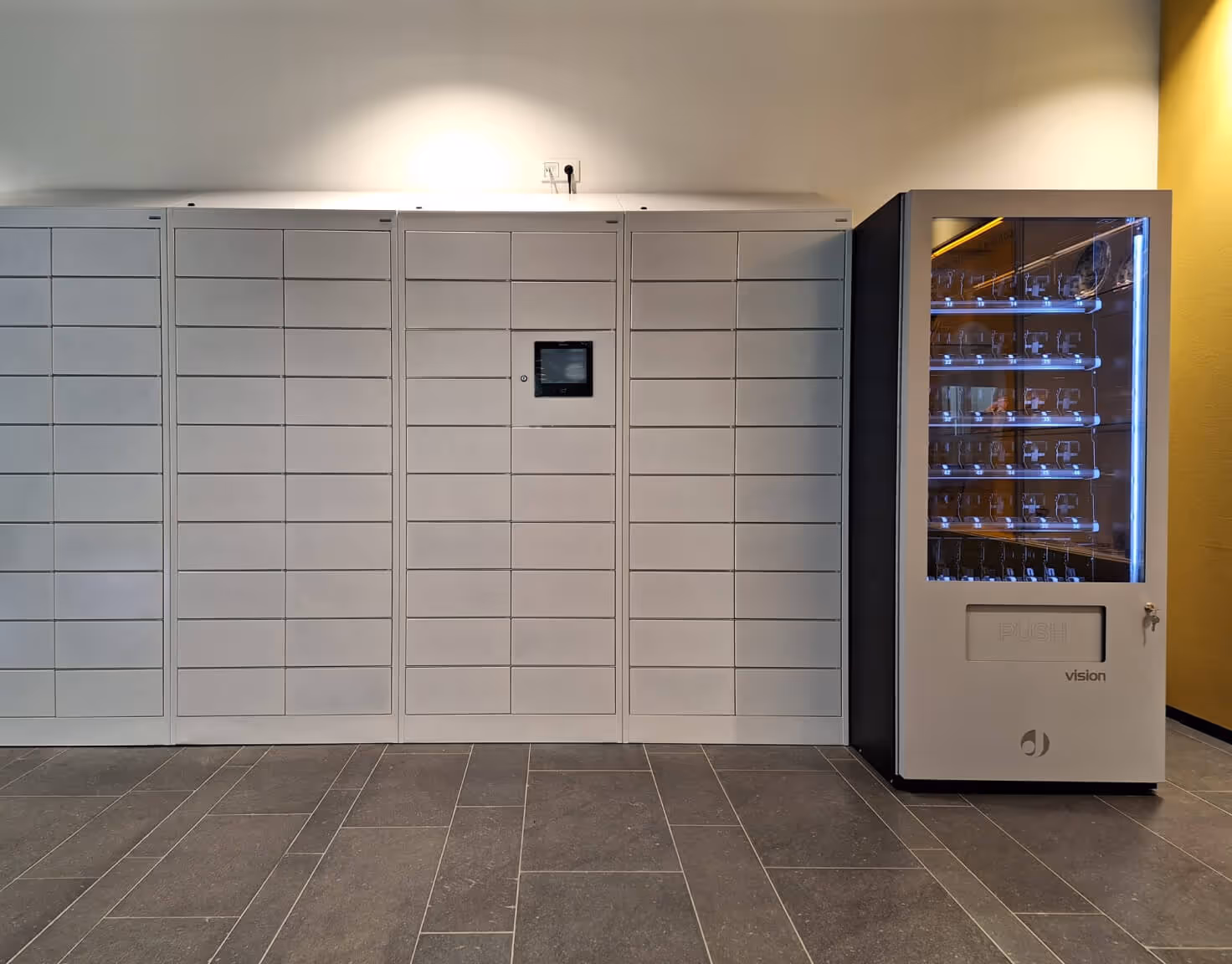 A set of white lockers with a central touchscreen panel next to a vending machine labeled 'vision' on a tiled floor.