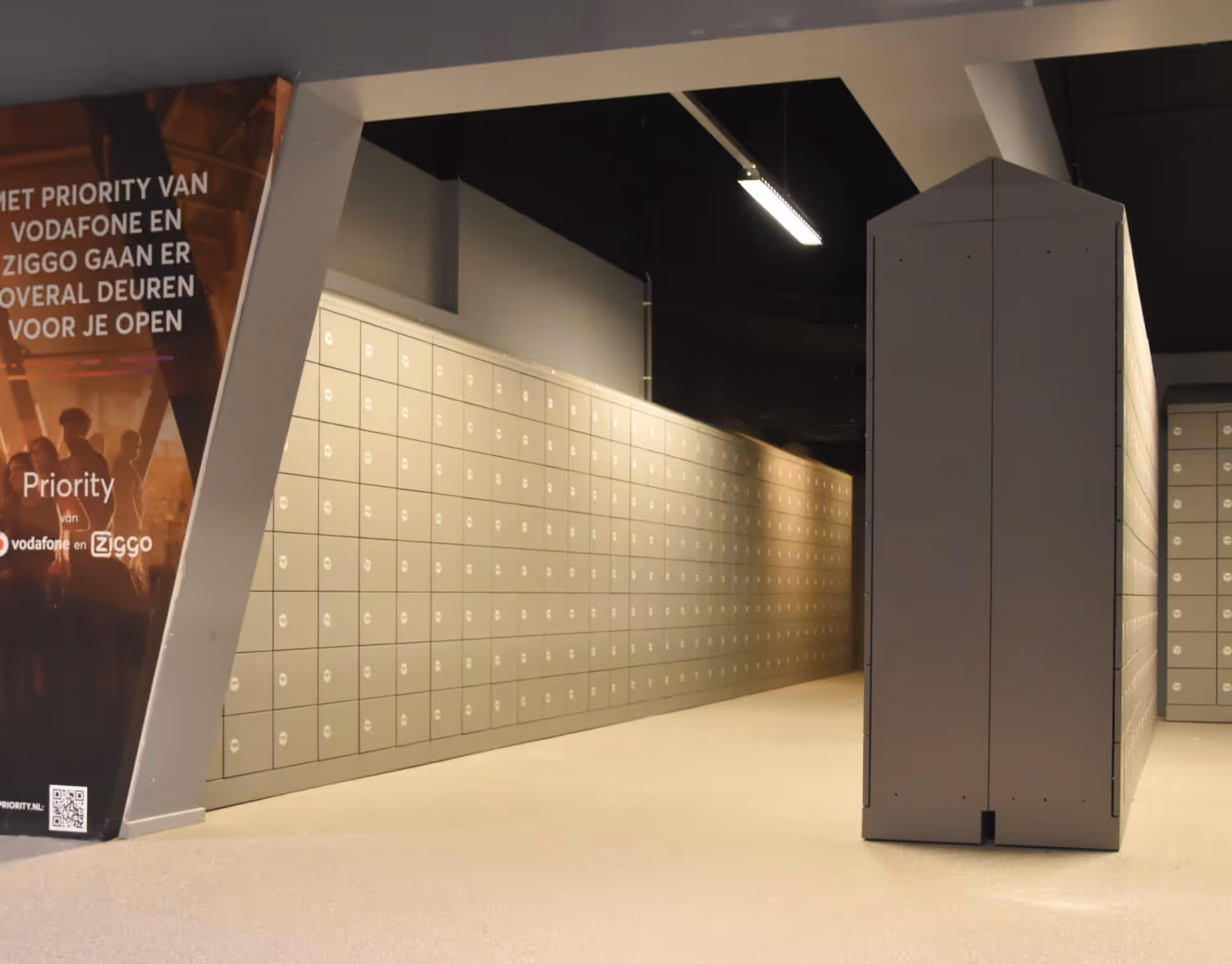Interior view of a room with rows of gray lockers and a large angled panel displaying a Vodafone Ziggo advertisement.