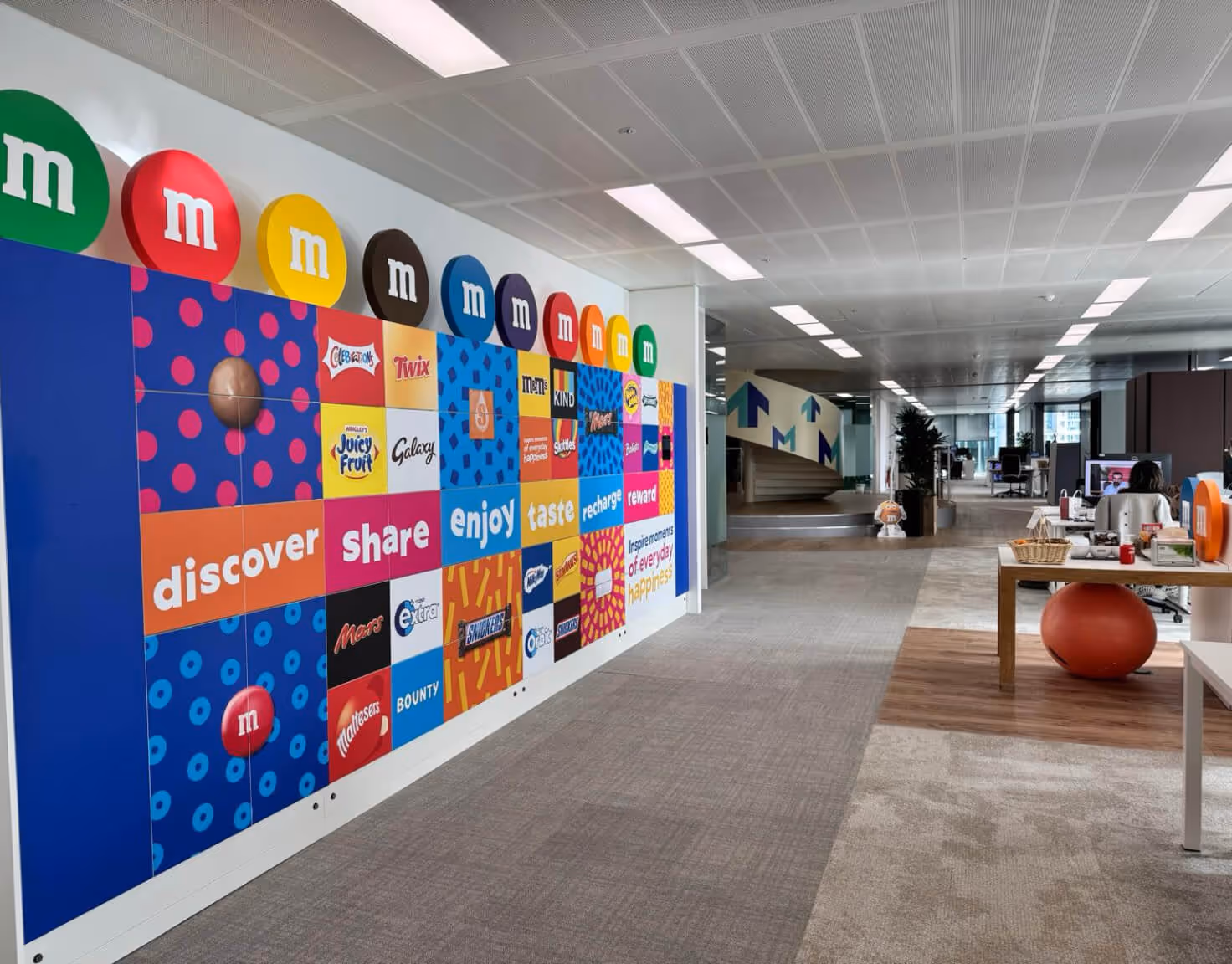 Modern office space with a colorful wall display featuring M&M's and other candy brand logos and slogans, alongside work desks and a staircase.