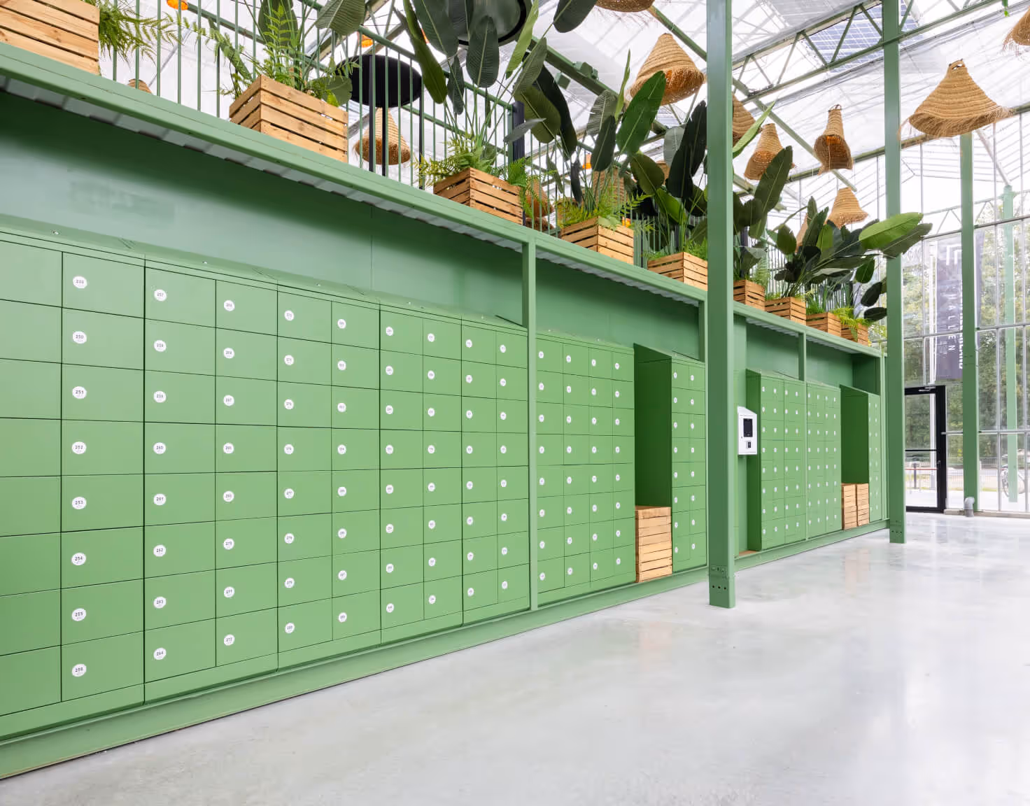 Modern green parcel lockers lined up inside a bright, glass-walled building with wooden planters containing tropical plants above.