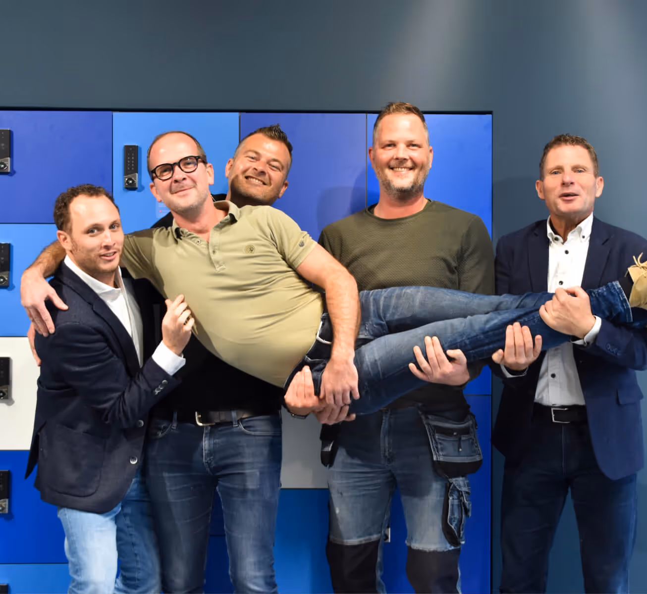 Four men are holding another man horizontally in front of blue lockers, all smiling and posing for the photo.
