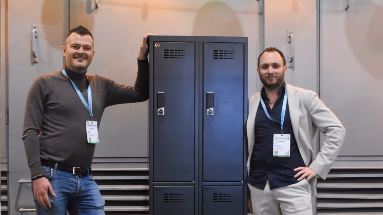 Two men standing on either side of a pair of dark lockers, smiling and wearing conference badges.