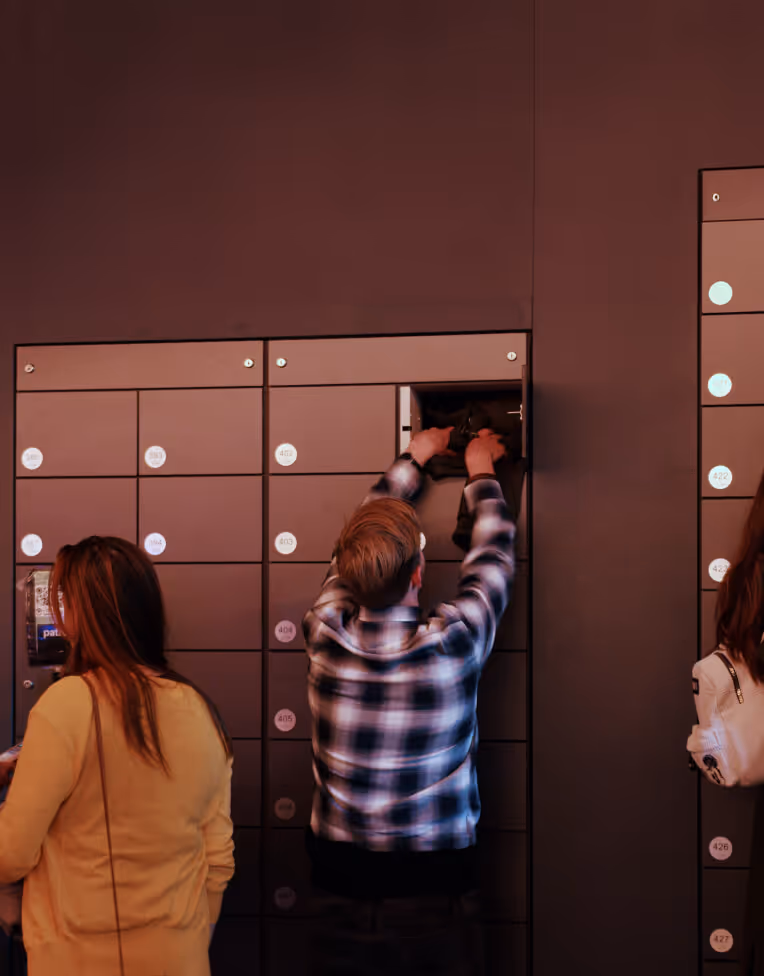 Person placing a bag into an open locker in a row of numbered lockers with two other people nearby.