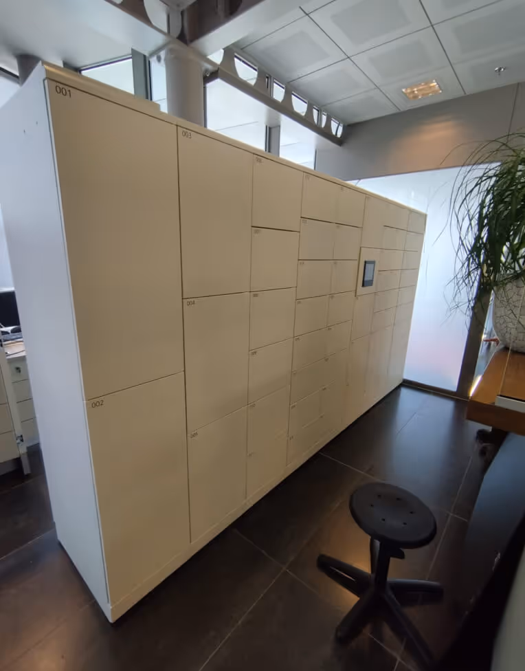 Beige parcel locker system with multiple numbered compartments in an office setting, next to a black swivel stool and a plant.