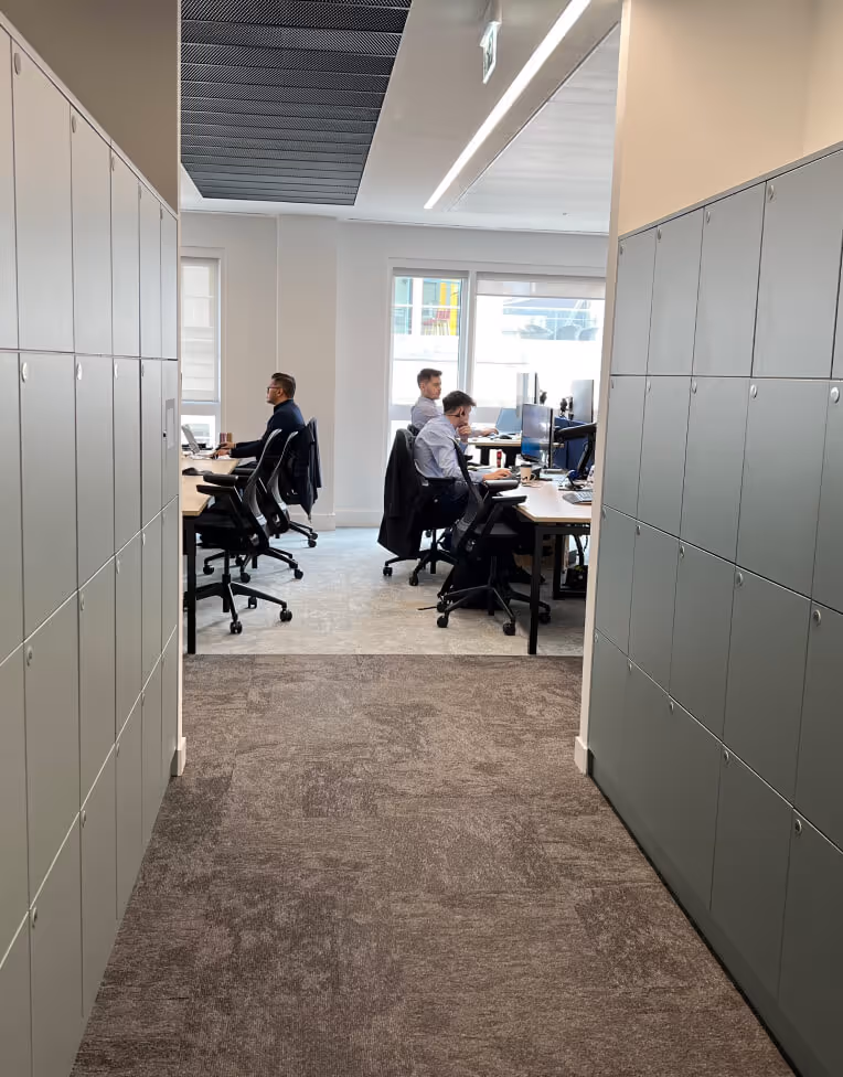 Office hallway with lockers on both sides leading to a workspace where three people are working at desks with computers.