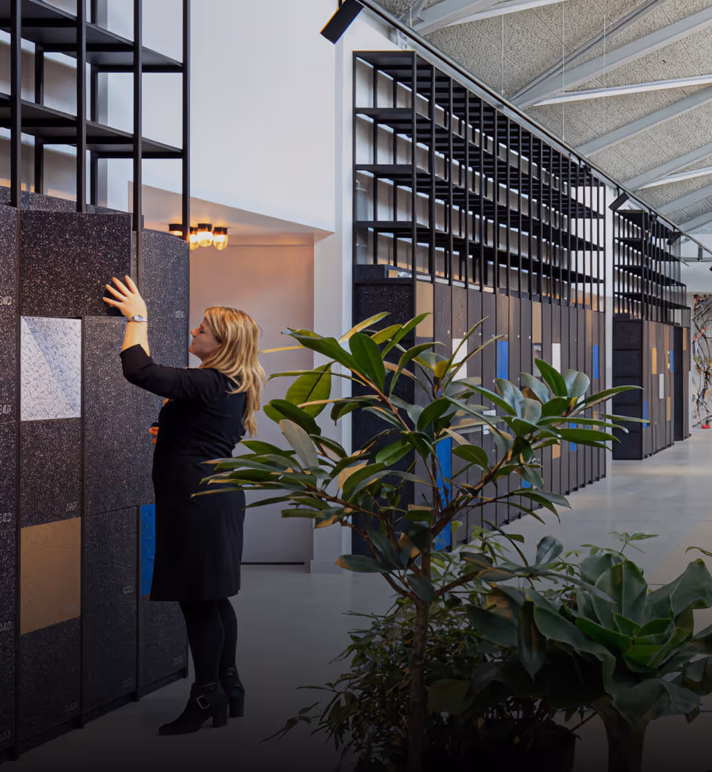 Woman examining large, vertical tile or stone samples displayed on black frames in a spacious, modern showroom with plants in the foreground.