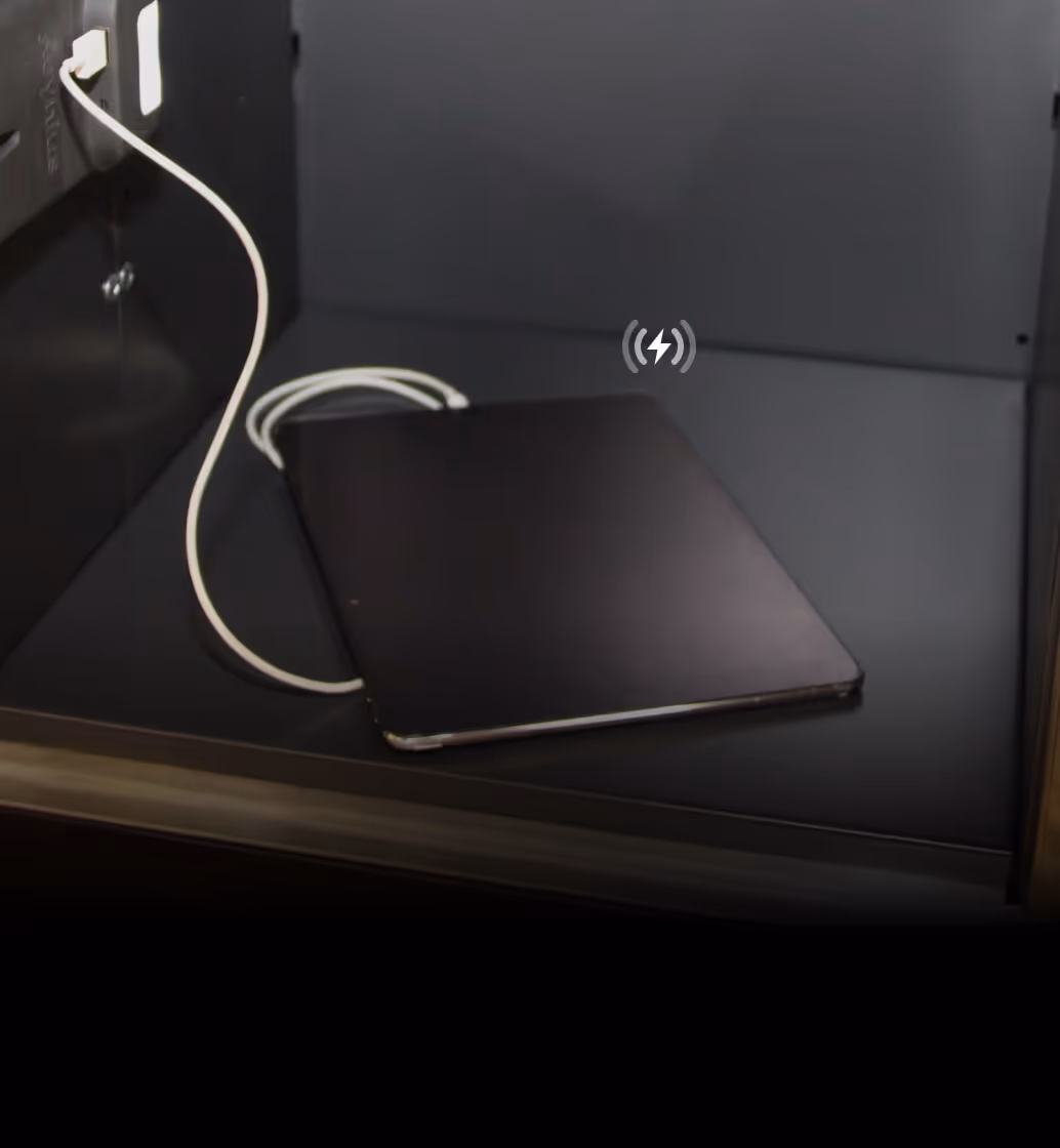 Closed black laptop charging inside a dark cubicle with a white power cable plugged into an outlet.