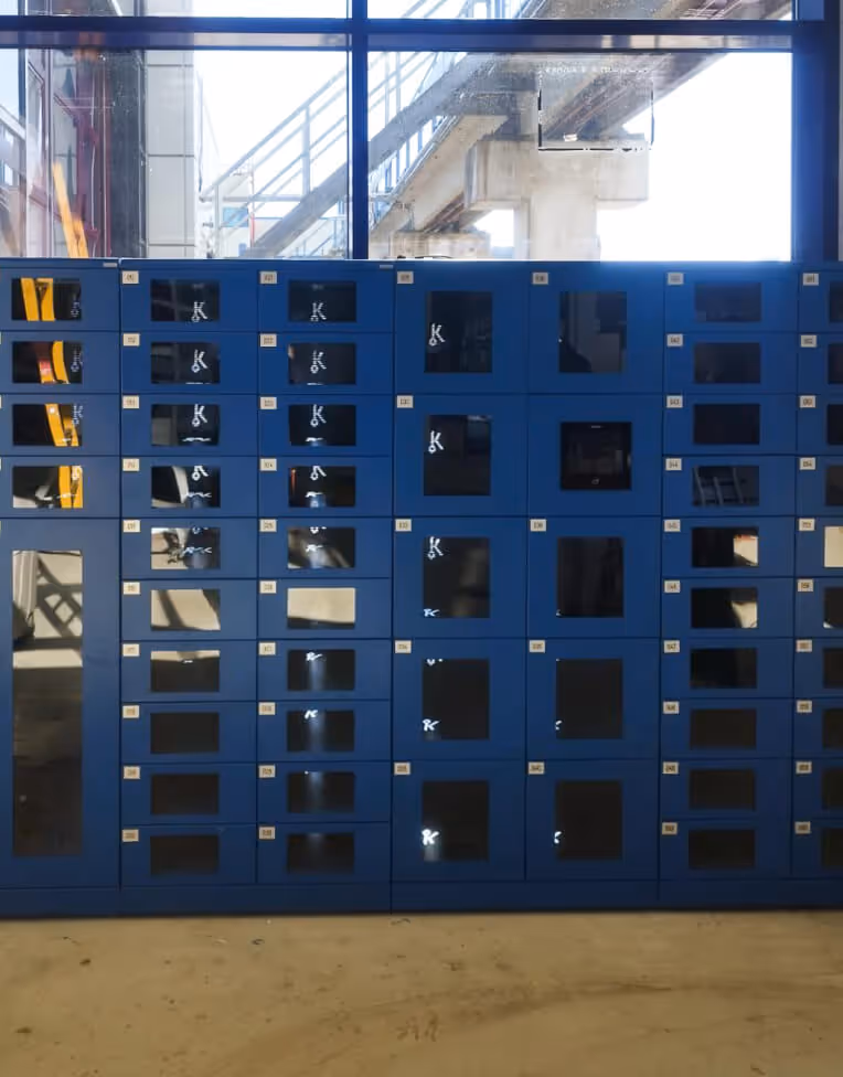 Rows of blue lockers with reflective glass doors inside a brightly lit room with large windows showing industrial structures outside.