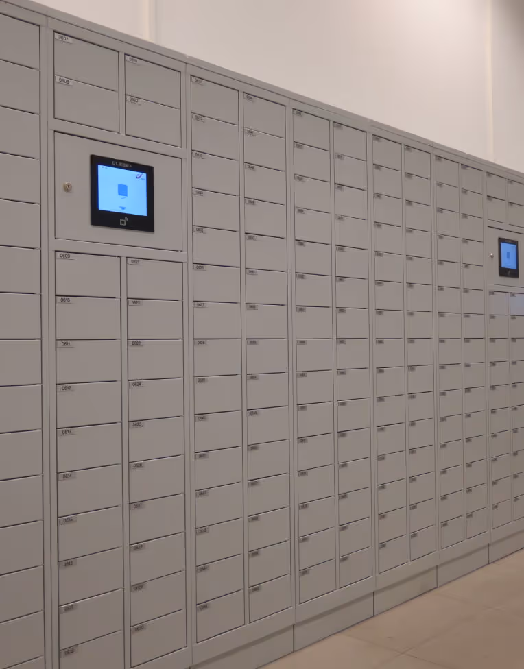 Row of gray electronic parcel lockers with digital screens in a well-lit indoor space.