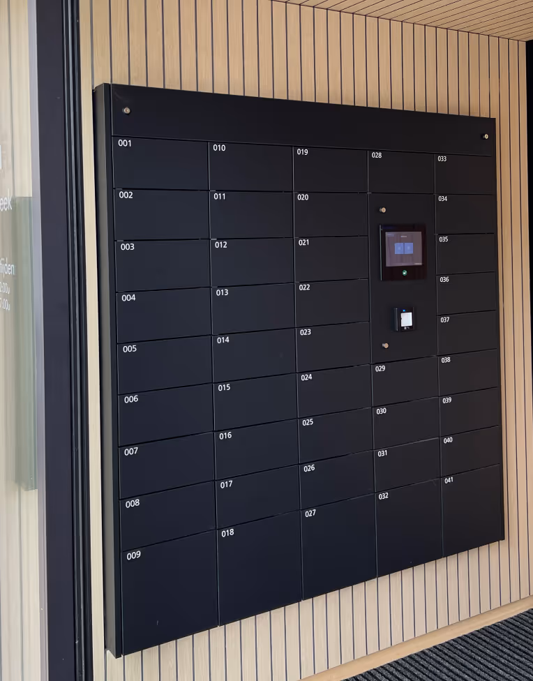 Black wall-mounted parcel locker system with numbered compartments and a digital touchscreen interface on a wooden slat wall.