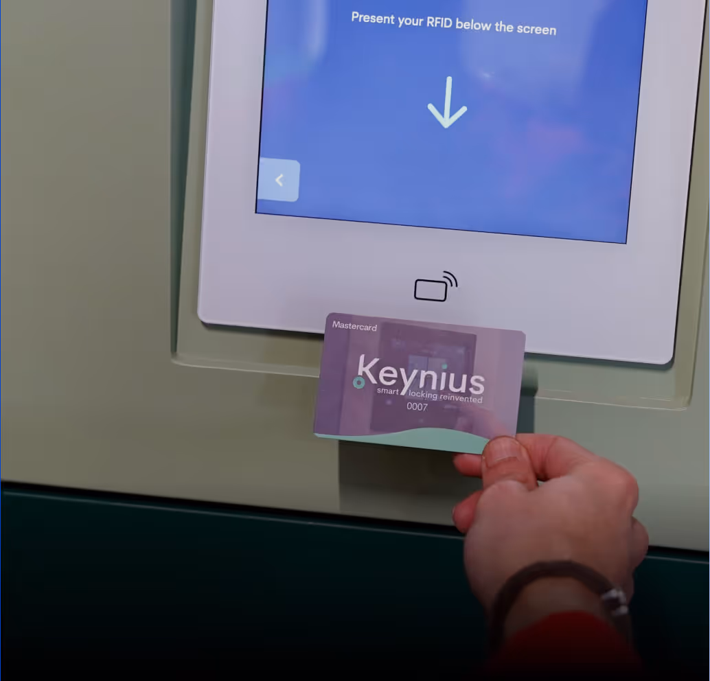 Person holding a Keynius RFID card in front of a vending machine screen that reads 'Present your RFID below the screen.'