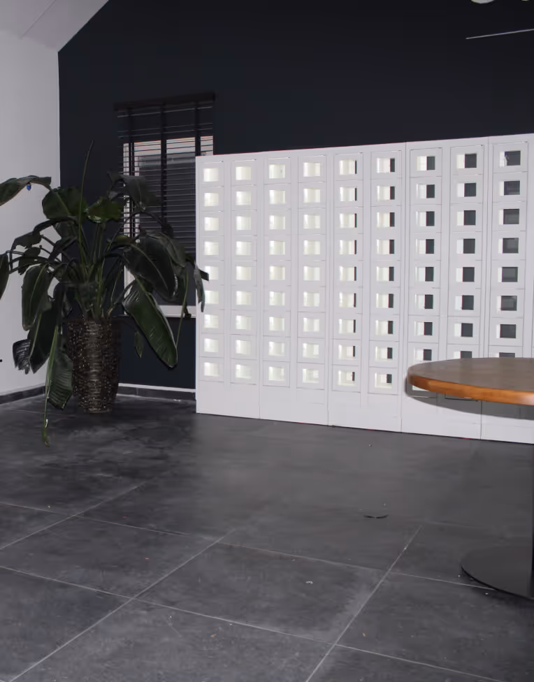 Modern interior with large green potted plant, dark gray tiled floor, white illuminated wall panels, and a round wooden table edge visible.