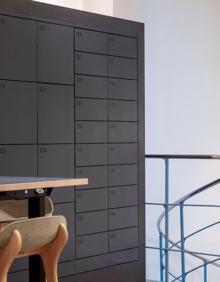 Modern interior featuring a wooden chair, a light wood desk, and a dark wall with numbered lockers beside a metal railing.