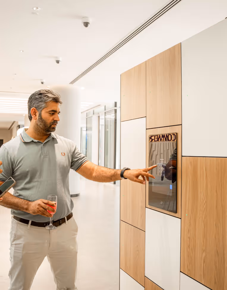 Man in casual clothes holding a glass and touching a wall-mounted digital screen in a modern office hallway.