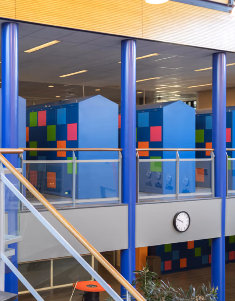 Interior of a modern building with blue pillars and colorful geometric panels on upper-level walls, a clock on the mid-level wall, and a staircase with wooden handrail.