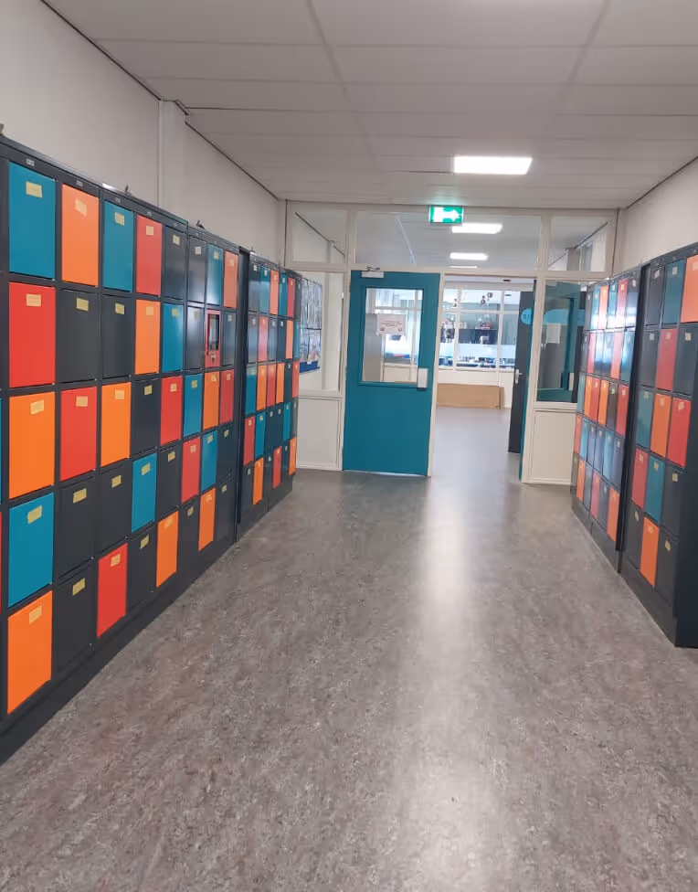 School hallway with lockers on both sides painted in teal, red, and orange, leading to a teal door with windows at the end.
