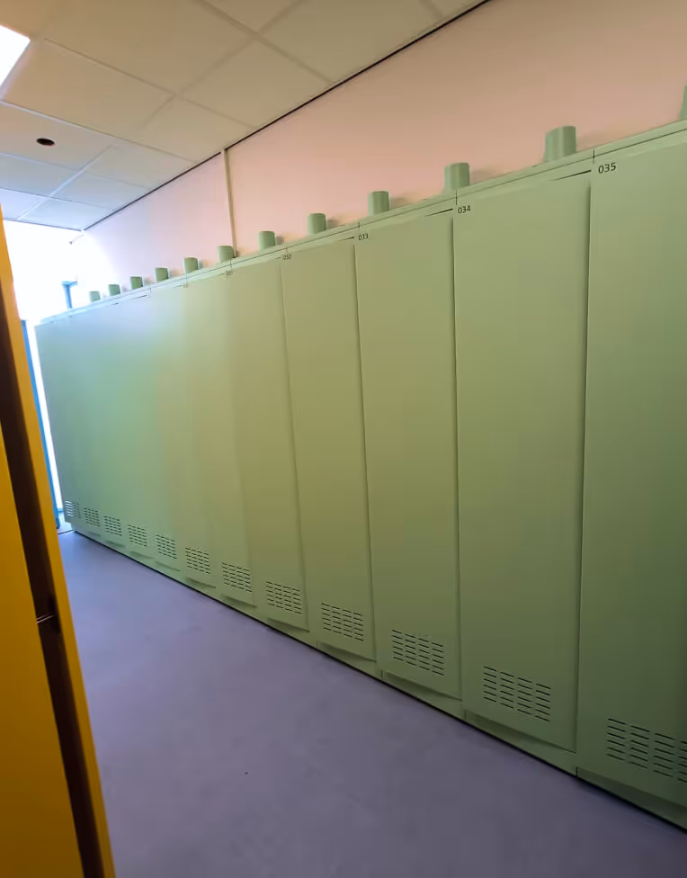 Row of green metal lockers with ventilation vents at the bottom in a hallway with a tiled ceiling and pinkish walls.