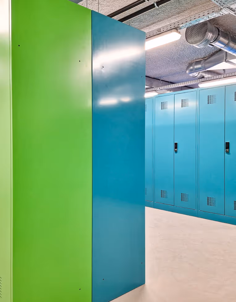 Row of green and blue lockers in a locker room with visible ceiling pipes and fluorescent lights.