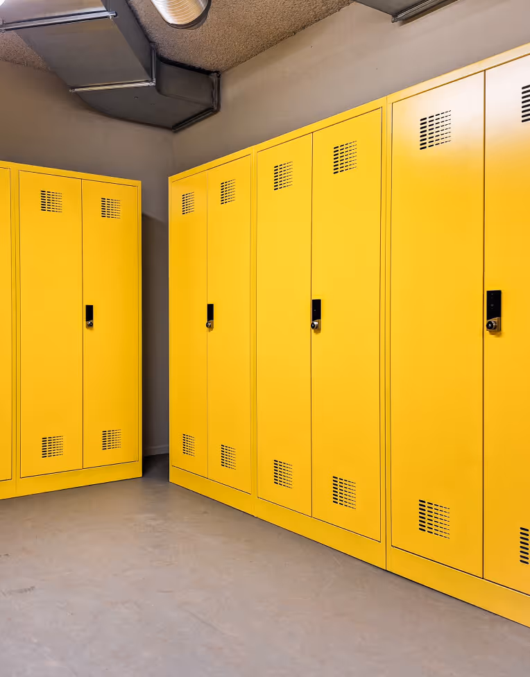 Row of bright yellow lockers with black handles in a clean room with gray floor and exposed ceiling ducts.