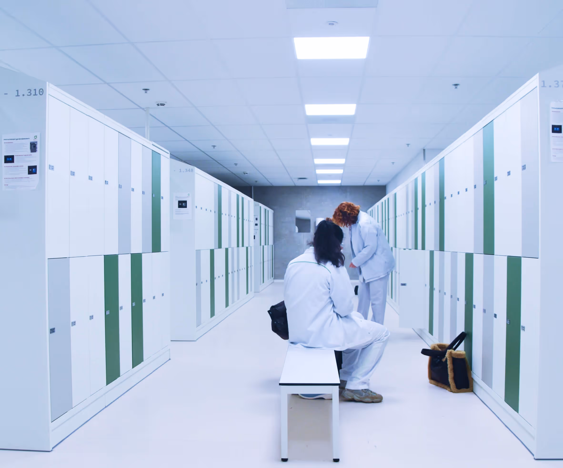 Two medical staff in white uniforms in a bright locker room with rows of white lockers with green accents.