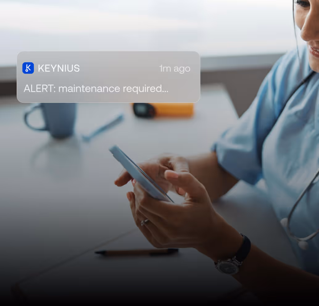 Person in light blue shirt holding a smartphone with a notification reading 'ALERT: maintenance required' from KEYNIUS.