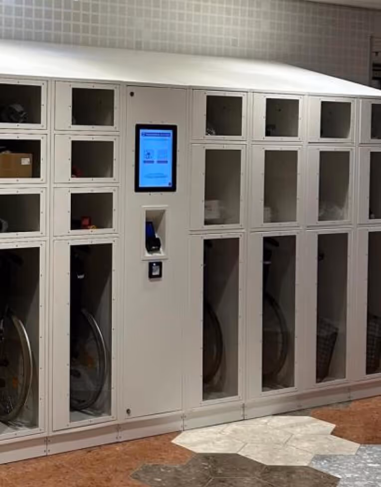 White laundry lockers with transparent doors and a digital touchscreen interface in the center, located indoors on a patterned floor.
